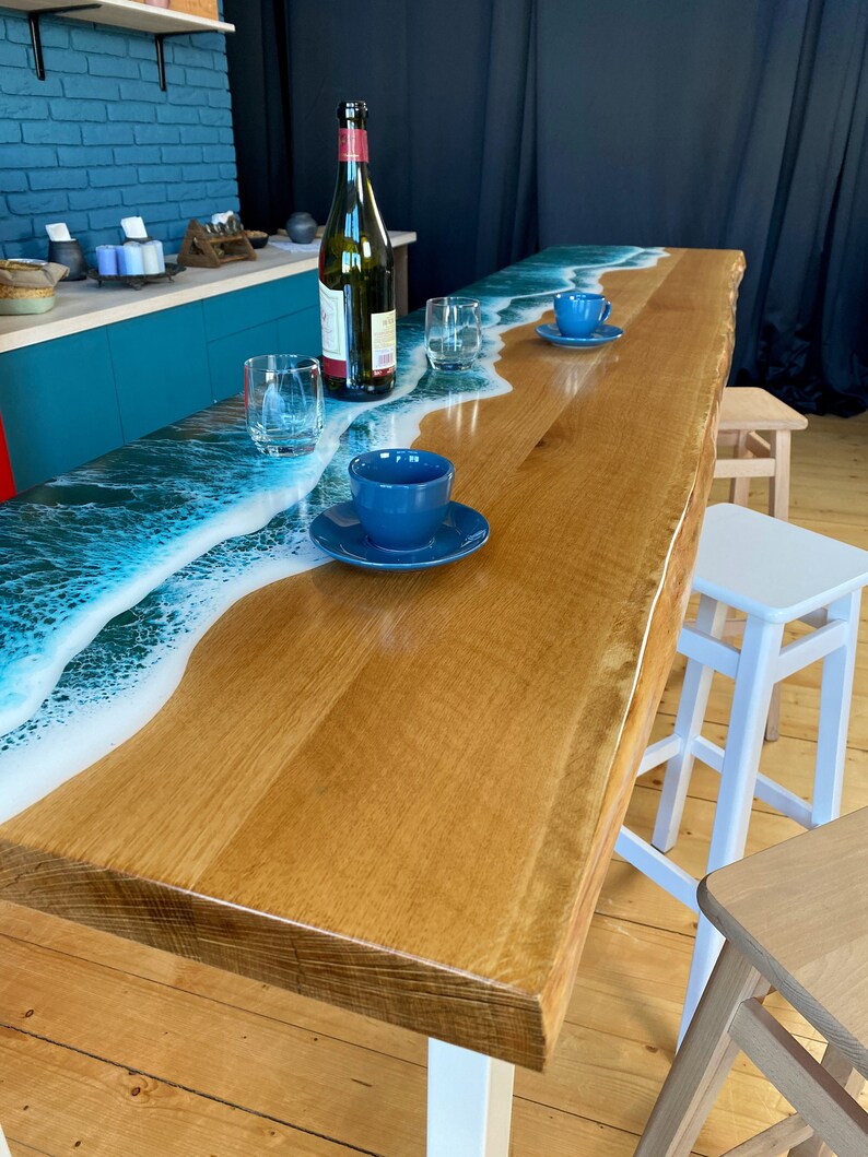 May include: A wooden table with a white epoxy resin top featuring a blue ocean wave design. The table has a white base and is surrounded by wooden and white stools.
