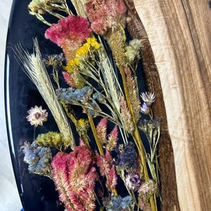 May include: A handcrafted resin art piece featuring dried flowers in shades of pink, yellow, and blue, set against a glossy black background. The artwork is partially bordered by natural wood, creating a unique contrast in textures and colors. A decorative item.
