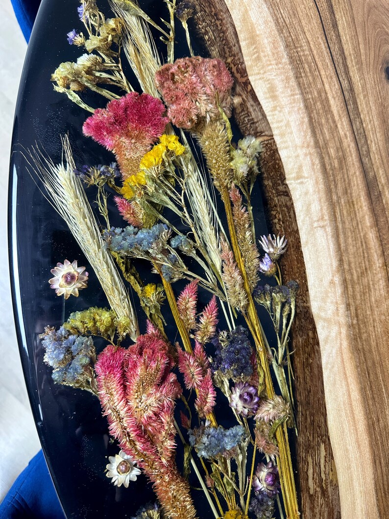 May include: A handcrafted resin art piece featuring dried flowers in shades of pink, yellow, and blue, set against a glossy black background. The artwork is partially bordered by natural wood, creating a unique contrast in textures and colors. A decorative item.