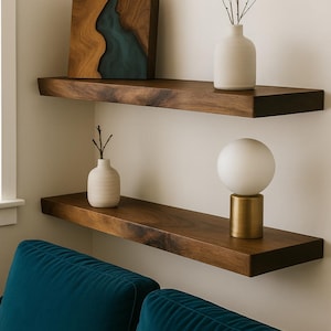 May include: Two dark wood floating shelves with decorative items. The top shelf holds a white vase with branches and a wooden art piece with a teal resin inlay. The bottom shelf has a small vase and a gold-based lamp with a white globe.