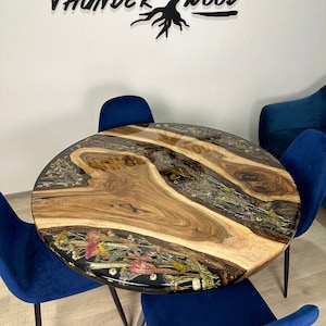 May include: Round wooden table with a live edge design, embedded with dried flowers and resin. The table is surrounded by four blue velvet chairs. The table's surface has a natural wood grain pattern. The wall behind the table has the text "THUNDER WOOD".