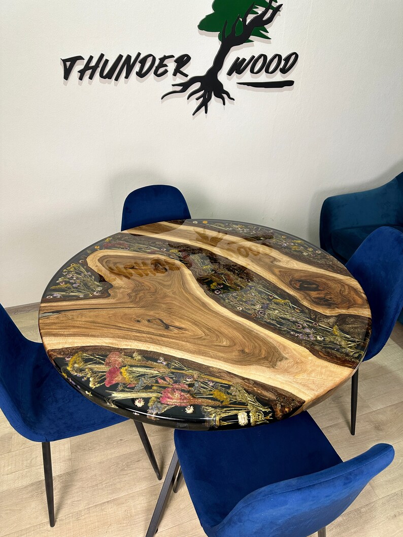 May include: Round wooden table with a live edge design, embedded with dried flowers and resin. The table is surrounded by four blue velvet chairs. The table's surface has a natural wood grain pattern. The wall behind the table has the text "THUNDER WOOD".