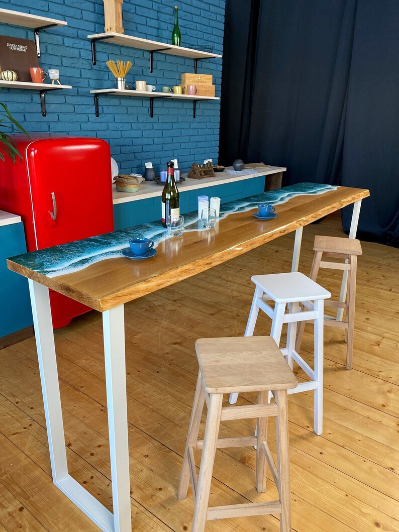 May include: A long wooden bar table with a blue and white epoxy resin wave design. The table has white metal legs and is set up with three wooden and white bar stools. The table is in a room with a blue brick wall and a red refrigerator.
