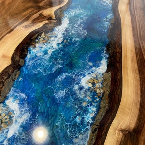 May include: A close-up of a wooden table top with a blue and white epoxy resin river design. The resin has a swirling pattern that resembles waves and includes white accents that look like foam.