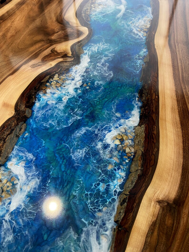 May include: A close-up of a wooden table top with a blue and white epoxy resin river design. The resin has a swirling pattern that resembles waves and includes white accents that look like foam.