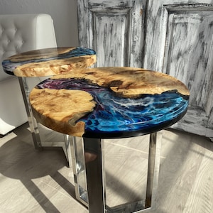 May include: Two round wooden end tables with a blue, white, and purple epoxy resin river design. The tables have a chrome metal base.