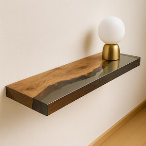 May include: A floating wooden shelf with a live edge design and a dark, glossy finish. The shelf is made of natural wood and has a clear, glass-like resin inlay. A small, white globe lamp with a gold base sits on the shelf, adding a touch of modern decor.