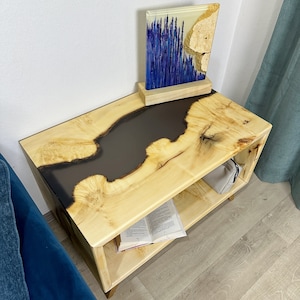 Puede incluir: Una mesa auxiliar de madera clara con un diseño de río de epoxi negro. La mesa tiene un estante inferior y está decorada con un libro y una obra de arte enmarcada con acentos morados y dorados.