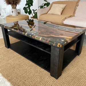 May include: A rectangular coffee table featuring a live edge wood slab centerpiece encased in clear resin. The resin border is embedded with colorful dried wildflowers. The table has a black wooden frame with a lower shelf and four sturdy legs.