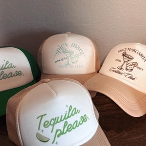 May include: Three trucker hats with different designs. The first hat is green and says "Tequila, please." The second hat is white with a green design and says "Spicy Margs, extra spicy Cocktail Club." The third hat is beige with a brown design and says "Spicy Margarita, extra spicy Cocktail Club."