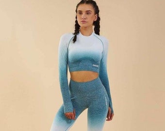 Könnte beinhalten: Eine Frau trägt ein langärmliges, ombrefarbenes, blau-weißes Crop-Top und eine passende Leggings. Das Top und die Leggings sind nahtlos und haben einen dezenten Glanz. Das Top hat den Schriftzug "GYMSHARK" auf der Vorderseite.