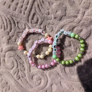 Może przedstawiać: Trzy bransoletki z koralików z białymi koralikami literowymi, które wymawiają słowa, przeplatane kolorowymi koralikami i zawieszkami w kształcie gwiazd. Jedna bransoletka jest różowa, jedna zielona, a jedna biała.