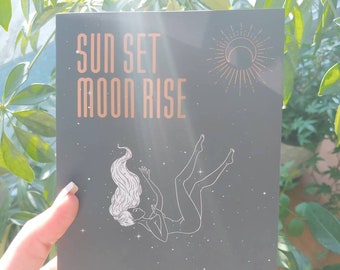 May include: A dark gray paperback book titled Sun Set Moon Rise by Maria Sayde Hraiki, featuring a white line art illustration of a person floating among stars, with copper-colored celestial sun and moon icons on the cover.