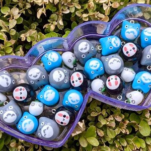 May include: A collection of small, colourful, hand-painted ceramic charms in a purple heart-shaped container. The charms feature various cartoon faces with different expressions. Some charms are blue, some are grey, and some are white.
