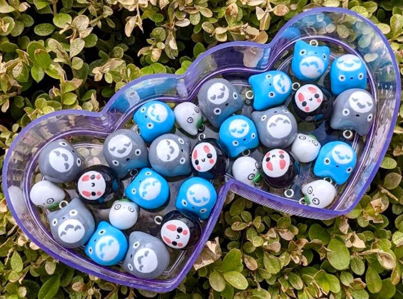 May include: A collection of small, colourful, hand-painted ceramic charms in a purple heart-shaped container. The charms feature various cartoon faces with different expressions. Some charms are blue, some are grey, and some are white.