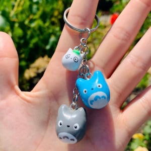 May include: A keychain featuring three small, hand-painted ceramic charms. The charms are shaped like the characters from the animated film My Neighbor Totoro. The charms are blue, grey, and white.