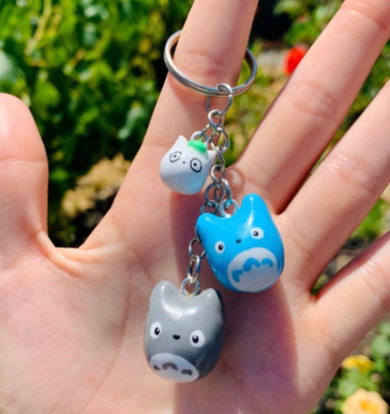 May include: A keychain featuring three small, hand-painted ceramic charms. The charms are shaped like the characters from the animated film My Neighbor Totoro. The charms are blue, grey, and white.