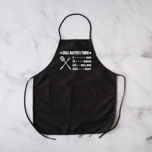 May include: Black grill master's apron with a white graphic of crossed grilling tools and a meat doneness guide. The guide includes the terms "Rare", "Medium", "Well Done", and "Crispy".