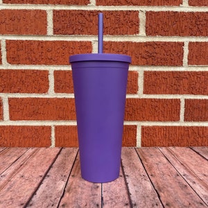 May include: A tall, purple, reusable tumbler with a lid and a straw. The tumbler is sitting on a wooden surface in front of a brick wall.