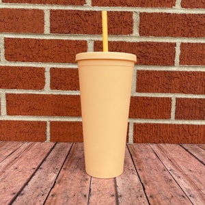 May include: A peach-colored reusable tumbler with a yellow straw. The tumbler is made of plastic and has a smooth, matte finish. It is sitting on a wooden surface.