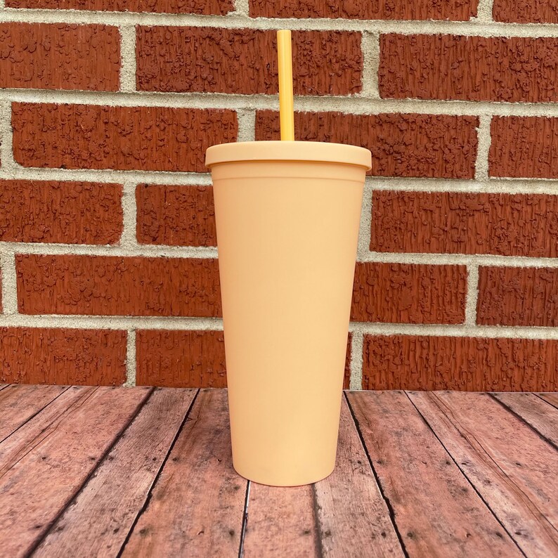 May include: A peach-colored reusable tumbler with a yellow straw. The tumbler is made of plastic and has a smooth, matte finish. It is sitting on a wooden surface.