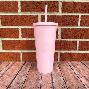 May include: A pink stainless steel tumbler with a lid and a straw. The tumbler is sitting on a wooden surface.