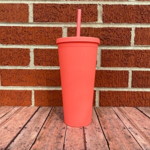 May include: A coral pink reusable tumbler with a lid and a straw. The tumbler is sitting on a wooden surface.