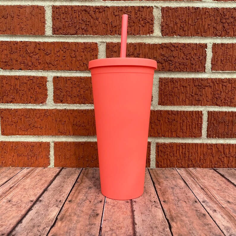 May include: A coral pink reusable tumbler with a lid and a straw. The tumbler is sitting on a wooden surface.