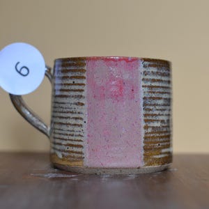 Può includere: Una tazza cilindrica in ceramica con un esterno marrone testurizzato e scanalato e una striscia verticale rosa liscia al centro. Un'etichetta circolare bianca con il numero nero 6 è attaccata al manico della tazza.