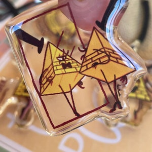 May include: A clear plastic keychain featuring a cartoon image of two yellow triangle-shaped characters with black eyes and red outlines. The characters are standing on their legs and have a black line connecting them. The keychain is on a brown surface.