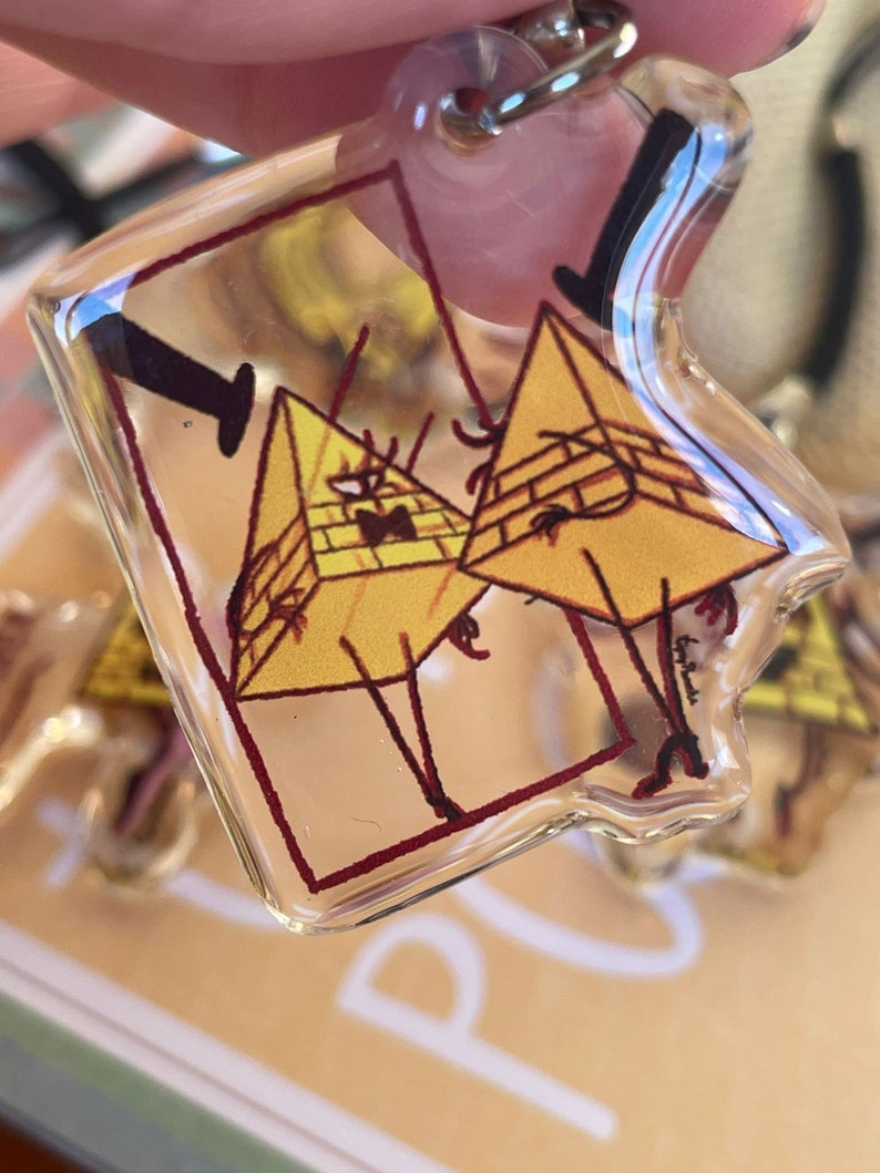 May include: A clear plastic keychain featuring a cartoon image of two yellow triangle-shaped characters with black eyes and red outlines. The characters are standing on their legs and have a black line connecting them. The keychain is on a brown surface.