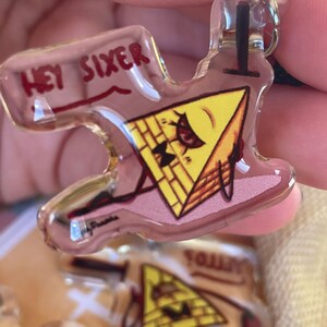 May include: A yellow triangle-shaped keychain with a cartoon face and the text "Hey Sixer" on the top. The keychain is made of clear acrylic and has a metal ring for attaching to a bag or keys.