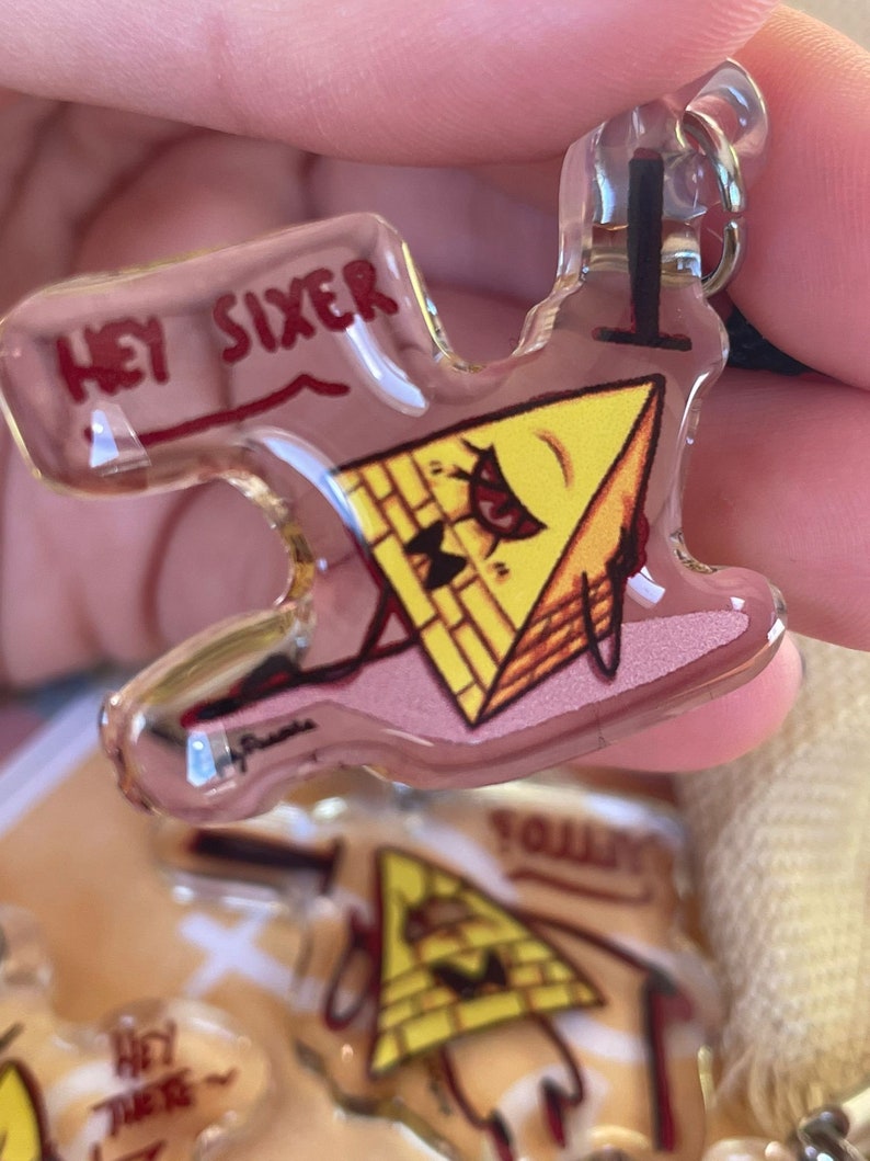 May include: A yellow triangle-shaped keychain with a cartoon face and the text "Hey Sixer" on the top. The keychain is made of clear acrylic and has a metal ring for attaching to a bag or keys.