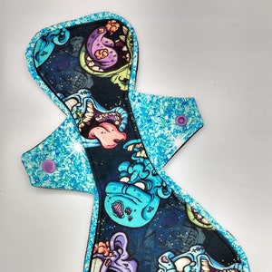 Pode incluir: Uma almofada menstrual de tecido reutilizável com um verso de glitter azul e uma frente de tecido preto com uma estampa de zumbis de desenho animado coloridos.