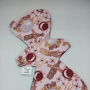 May include: Pink reusable cloth menstrual pad with a repeating pattern of hands, moons, stars, flowers, and bundles of sage. The text "Support Your Local Witch" is repeated throughout the pattern. The pad has a black border and a white label that says "Hand Made".