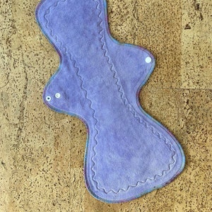 May include: A purple reusable cloth menstrual pad with a zig-zag stitched edge and white poppers.
