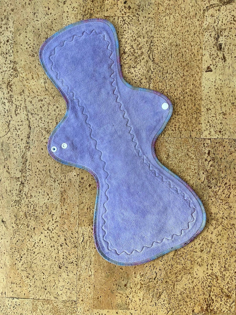 May include: A purple reusable cloth menstrual pad with a zig-zag stitched edge and white poppers.