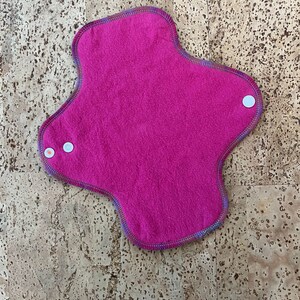 May include: A pink reusable cloth menstrual pad with a colourful trim and white poppers.