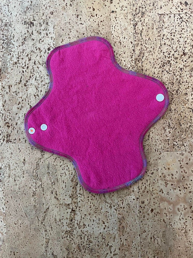 May include: A pink reusable cloth menstrual pad with a colourful trim and white poppers.