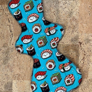 May include: A blue reusable cloth menstrual pad with a sushi print. The pad features various types of sushi, including maki, nigiri, and sashimi, all with cute faces.