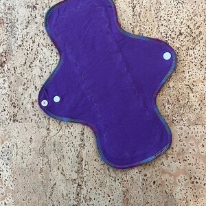 May include: Purple reusable cloth menstrual pad with white snap buttons on the sides.