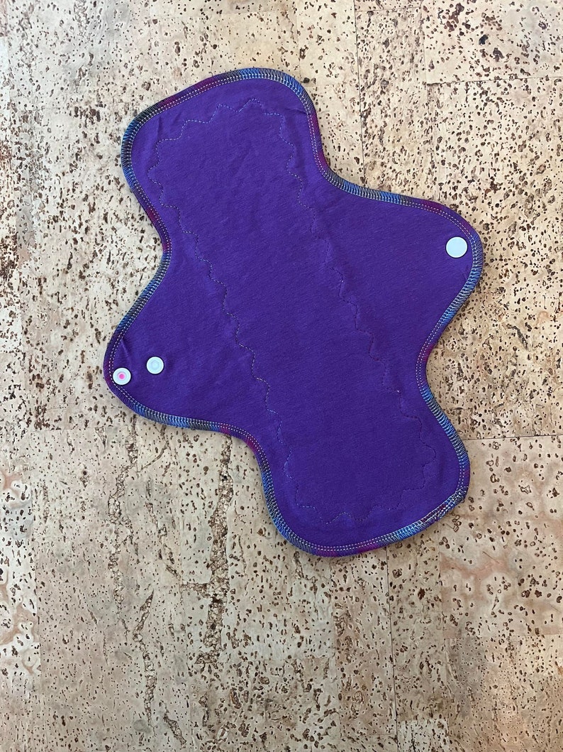 May include: Purple reusable cloth menstrual pad with white snap buttons on the sides.