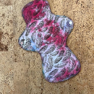 May include: A reusable cloth menstrual pad with a tie-dye pattern featuring a feather design in pink, blue, and white.