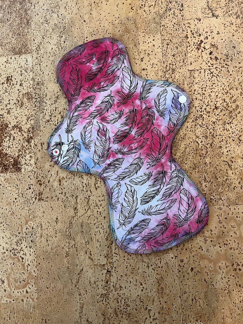 May include: A reusable cloth menstrual pad with a tie-dye pattern featuring a feather design in pink, blue, and white.