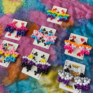 May include: A collection of beaded earrings in various pride flag colors, including rainbow, bisexual, transgender, lesbian, non-binary, and asexual designs. Each pair is displayed on a playing card, showcasing the unique color combinations and patterns.