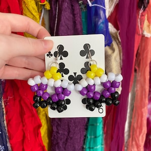 May include: A pair of star-shaped earrings crafted from plastic pony beads in yellow, white, purple, and black. The earrings are attached to a white card featuring a black clover suit pattern from a playing card, suspended by silver metal hooks.