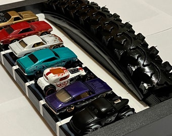May include: A black tire track with a row of six toy cars in different colors, including red, white, blue, green, and black. The cars are lined up on a black and white checkered surface.