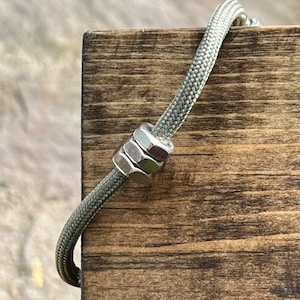 Könnte beinhalten: Ein grünes Paracord-Armband mit einer silbernen Metallperle. Die Perle besteht aus übereinander gestapelten Unterlegscheiben.