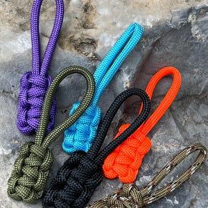 May include: Six zipper pulls made of woven paracord in purple, olive green, bright blue, black, neon orange, and a camouflage pattern. Each pull features a knotted rectangular base with a looped top for attachment to zippers or gear.