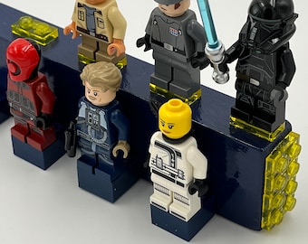 Puede incluir: Una colección de seis minifiguras, incluyendo una figura roja y negra, una figura azul y gris, una figura blanca y negra, una figura marrón y beige, una figura gris con una espada láser azul y una figura negra con un casco negro. Las figuras están de pie sobre bloques de Lego azules y amarillos.